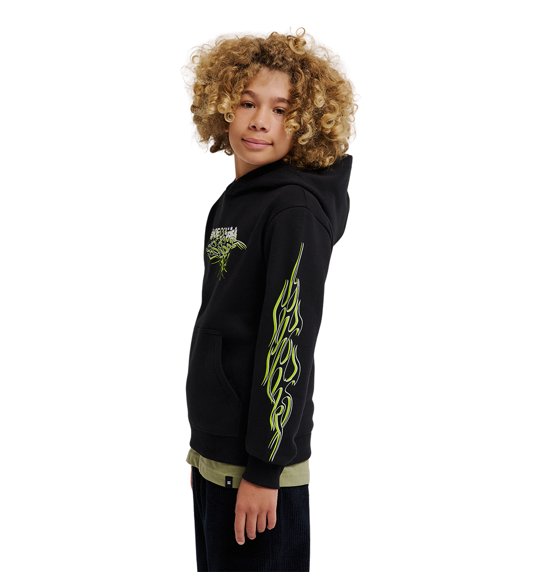 A young person with curly hair wears the DC Shoes Side Flame Hoodie Youth, a black fleece pullover featuring neon yellow-green DC graphics on the sleeve and chest, standing against a plain white background.
