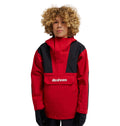 A person with curly blond hair wears the DC Shoes DC-43 Youth Anorak, a red and black insulated snow jacket featuring the “dcshoes” logo on the front pocket, standing against a plain white background.