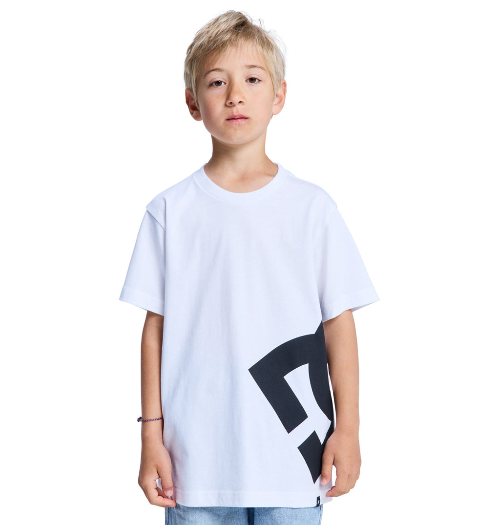 A young boy with blonde hair wears a Reebok Lanai Youth Hss white t-shirt featuring a bold black graphic on the lower side, standing against a plain white background.