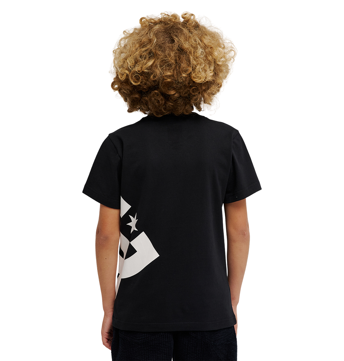 A child with curly blond hair faces away from the camera, wearing the DC Shoes DC Lanai Tee Youth—a relaxed fit black skate tee with a partial white graphic on the lower left—and black pants, standing against a plain white background.