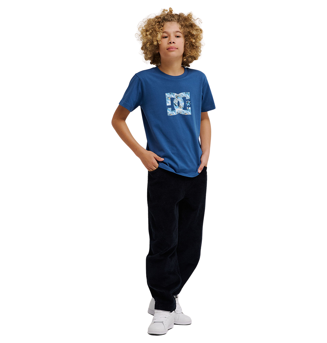 A young person with curly blond hair wears the DC Shoes DC Star Filled Tee Youth—a relaxed-fit blue graphic skate t-shirt with bold star artwork—paired with black pants and white sneakers, standing confidently with one hand in their pocket against a plain white background.