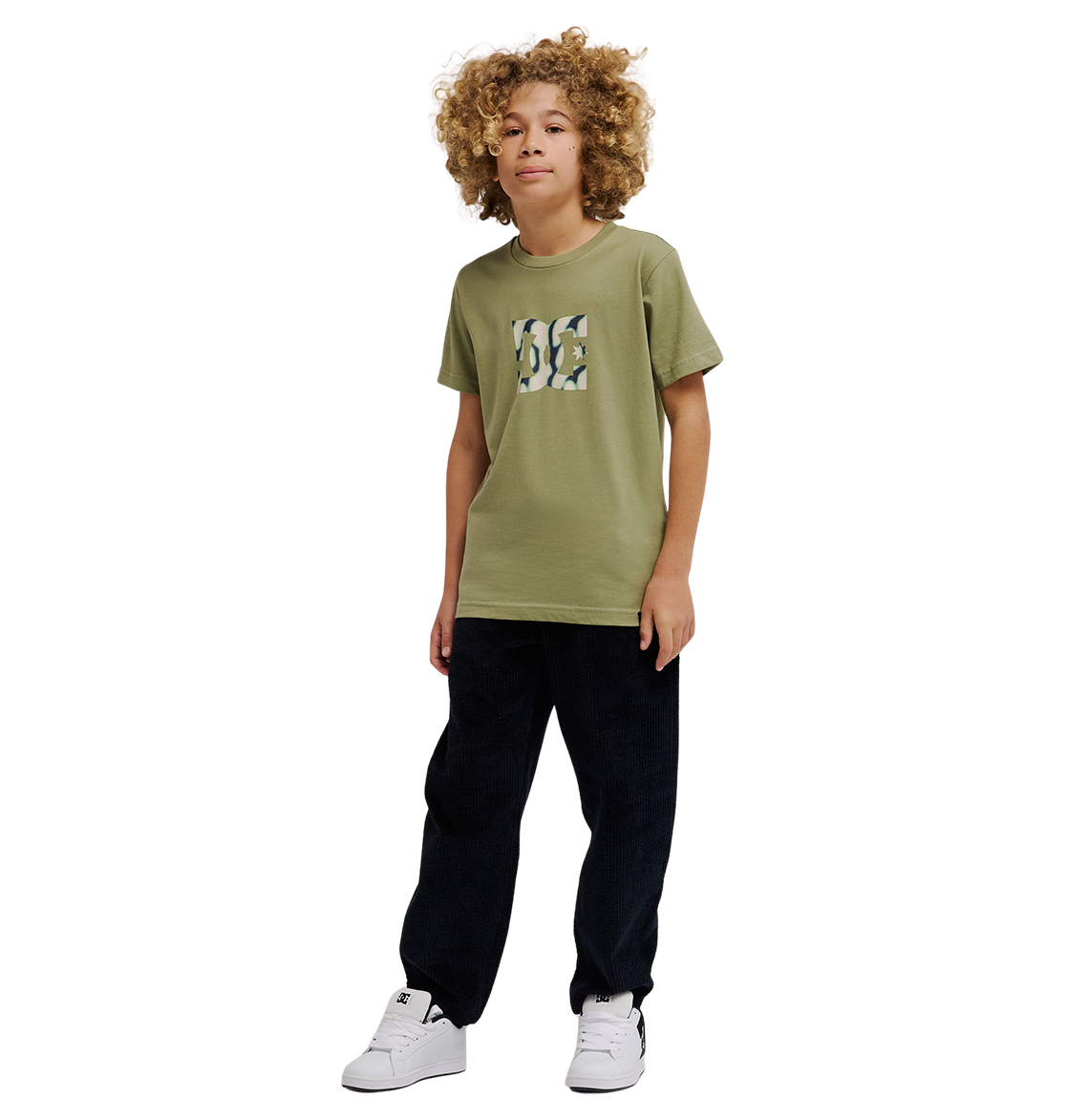 A young person with curly hair wears the DC Shoes DC Star Filled Tee Youth in olive green—a relaxed fit skate t-shirt featuring the iconic DC star graphic—paired with black pants and white sneakers, standing against a plain white background.