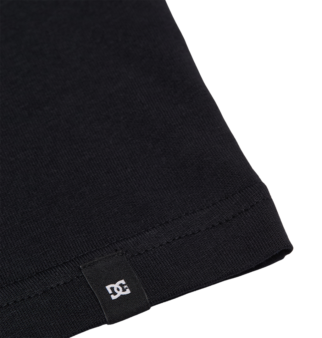 Close-up of the hem of the DC Star Filled Tee Youth, a black graphic skate tee by DC Shoes with a relaxed fit. A small black tag displaying the white DC Shoes logo is visible, set against a plain white background.