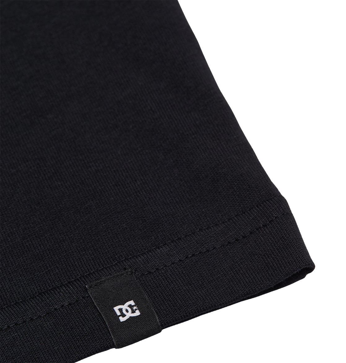 Close-up of the hem of the DC Star Filled Tee Youth, a black graphic skate tee by DC Shoes with a relaxed fit. A small black tag displaying the white DC Shoes logo is visible, set against a plain white background.