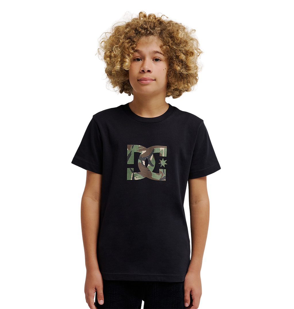 A young person with curly blond hair wears the DC Shoes DC Star Filled Tee Youth, a relaxed fit black graphic skate T-shirt featuring a camo-print DC Shoes star artwork logo on the chest, standing against a plain white background.