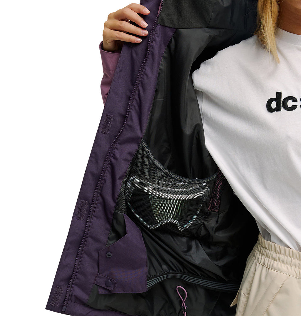 A person holds open a purple DC Shoes Everfrost Jacket, revealing an inner mesh pocket with goggles. The person wears a white shirt with black text and light-colored pants.