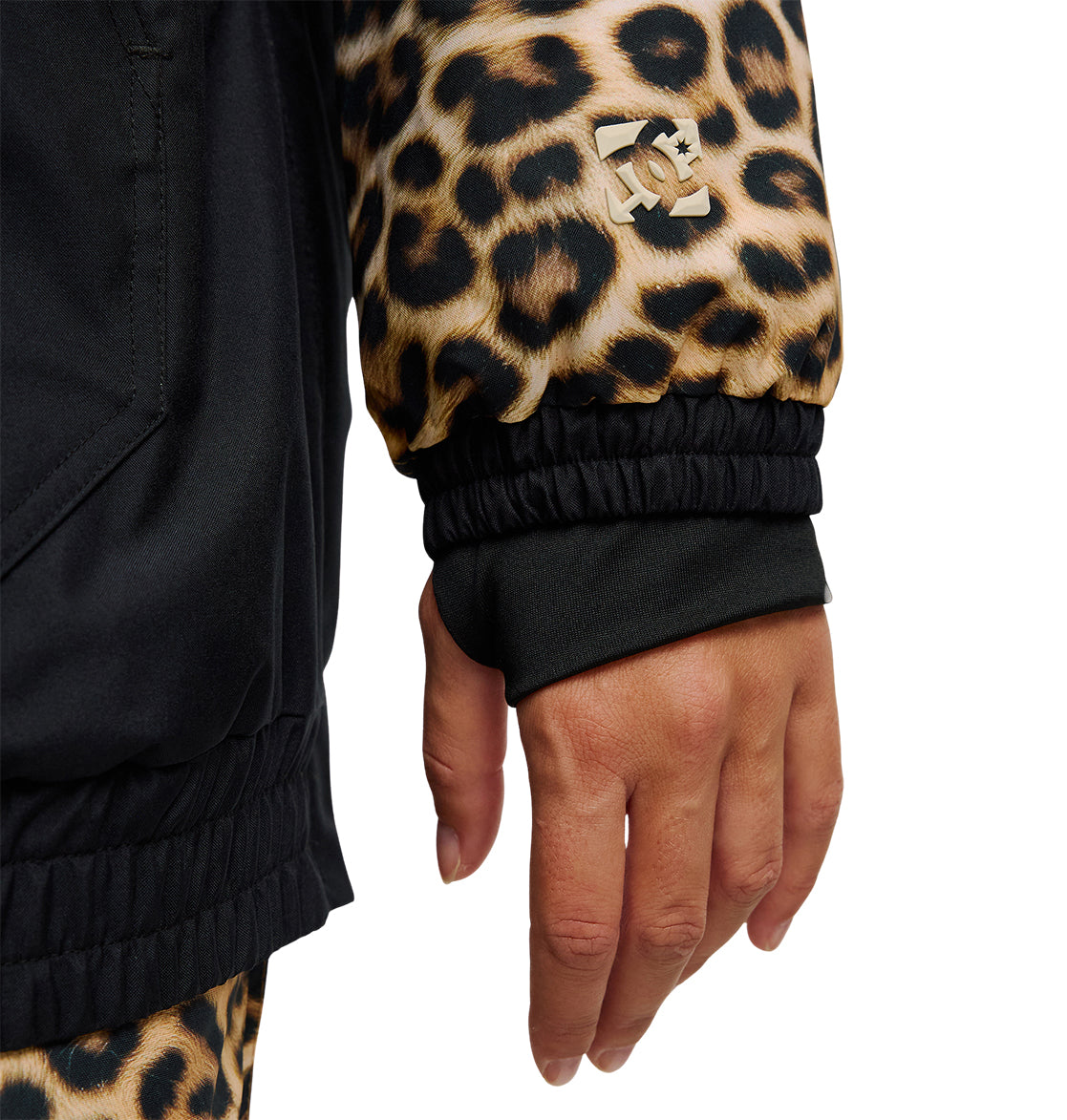 A hand is shown wearing the DC Shoes Chalet Anorak, a black insulated jacket with a leopard-print sleeve featuring a gold DC logo. Partially visible are matching leopard-print pants, designed for snow performance and enhanced waterproofing.