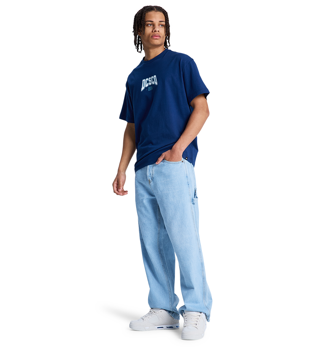 A person wears a navy DC Shoes t-shirt, white sneakers, and DC Shoes Men's Baggy Denim Carpenter Pant in a wide-leg fit, standing against a plain white background.