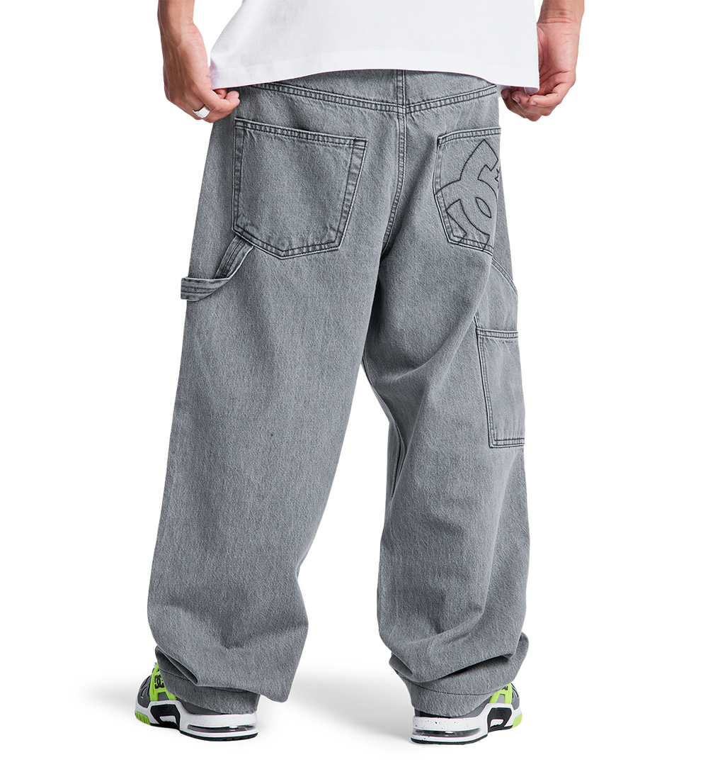 A person stands facing away, wearing DC Shoes Men's Baggy Denim Carpenter Pant with large pockets and a white graphic on the back pocket, styled with a white shirt and chunky sneakers featuring neon green accents.