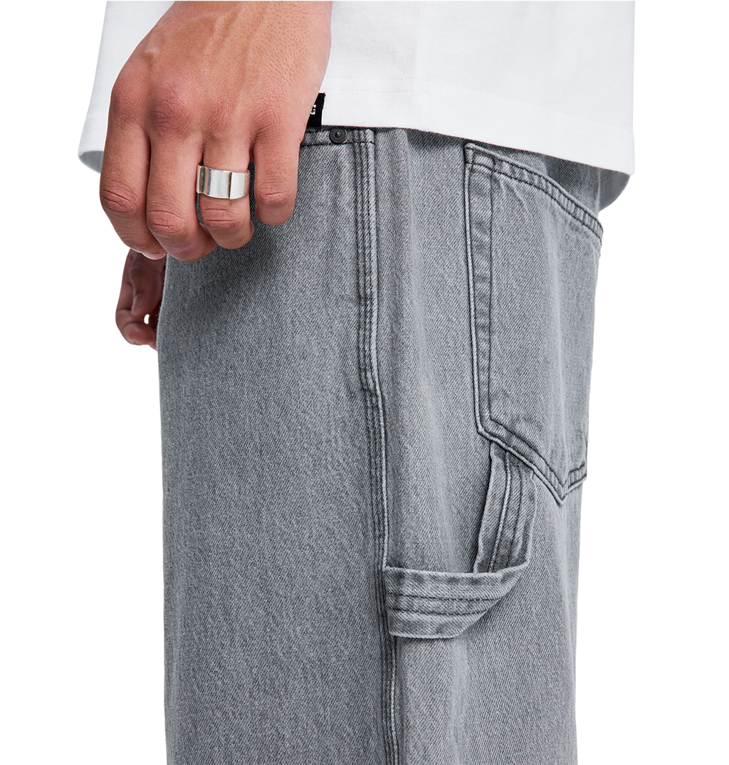 A close-up of a hand wearing a silver ring rests near the pocket of DC Shoes Men's Baggy Denim Carpenter Pant in light gray with visible stitching, shown next to a white shirt.