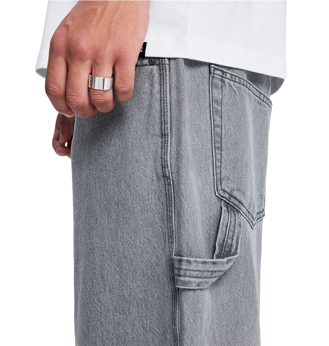 A close-up of a hand wearing a silver ring rests near the pocket of DC Shoes Men's Baggy Denim Carpenter Pant in light gray with visible stitching, shown next to a white shirt.