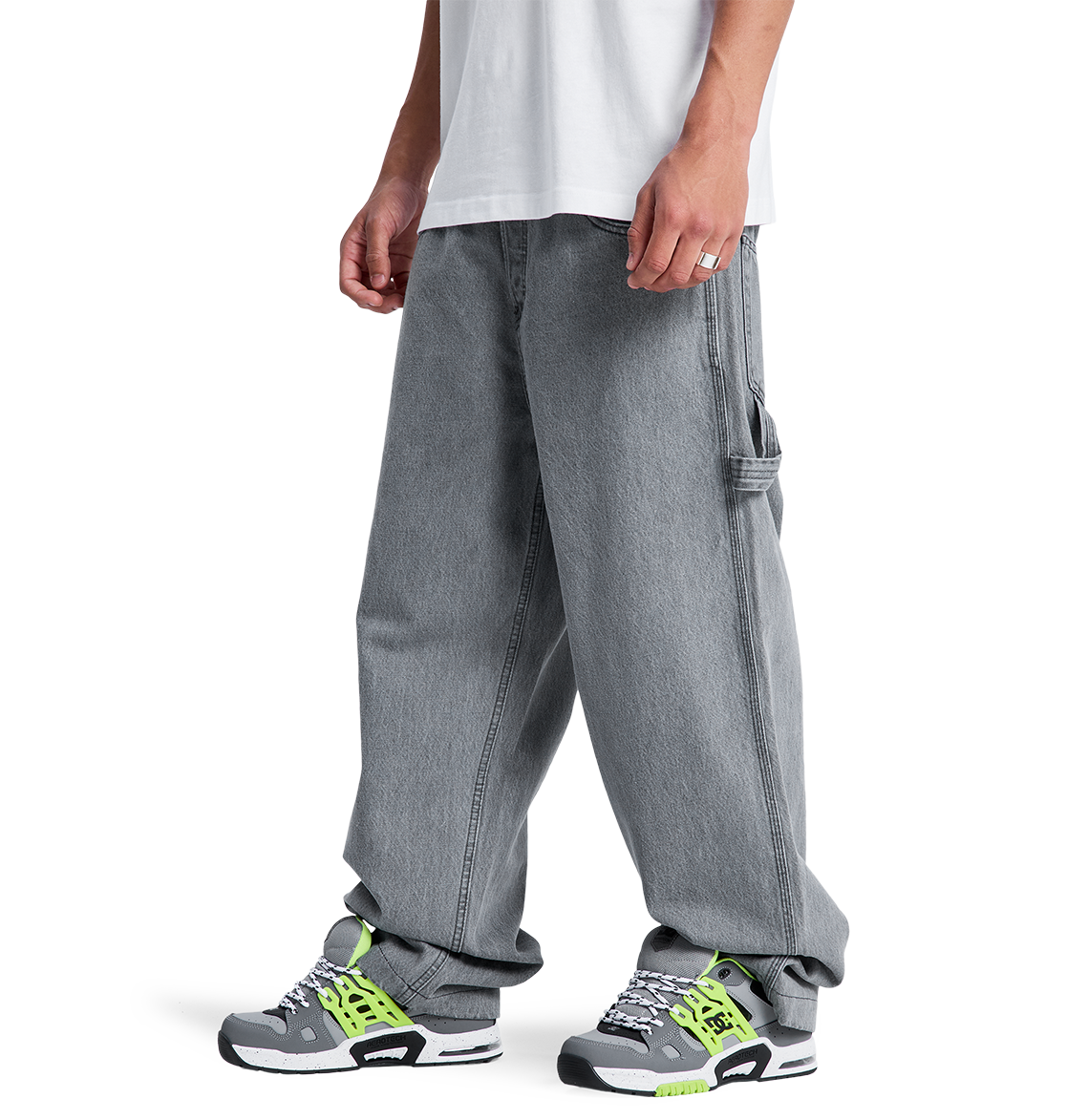 A person wears a white T-shirt, gray sneakers with white, black, and neon green accents, and DC Shoes Men's Baggy Denim Carpenter Pant—a wide-leg fit featuring heavyweight denim and side pocket detail that covers part of the sneakers.