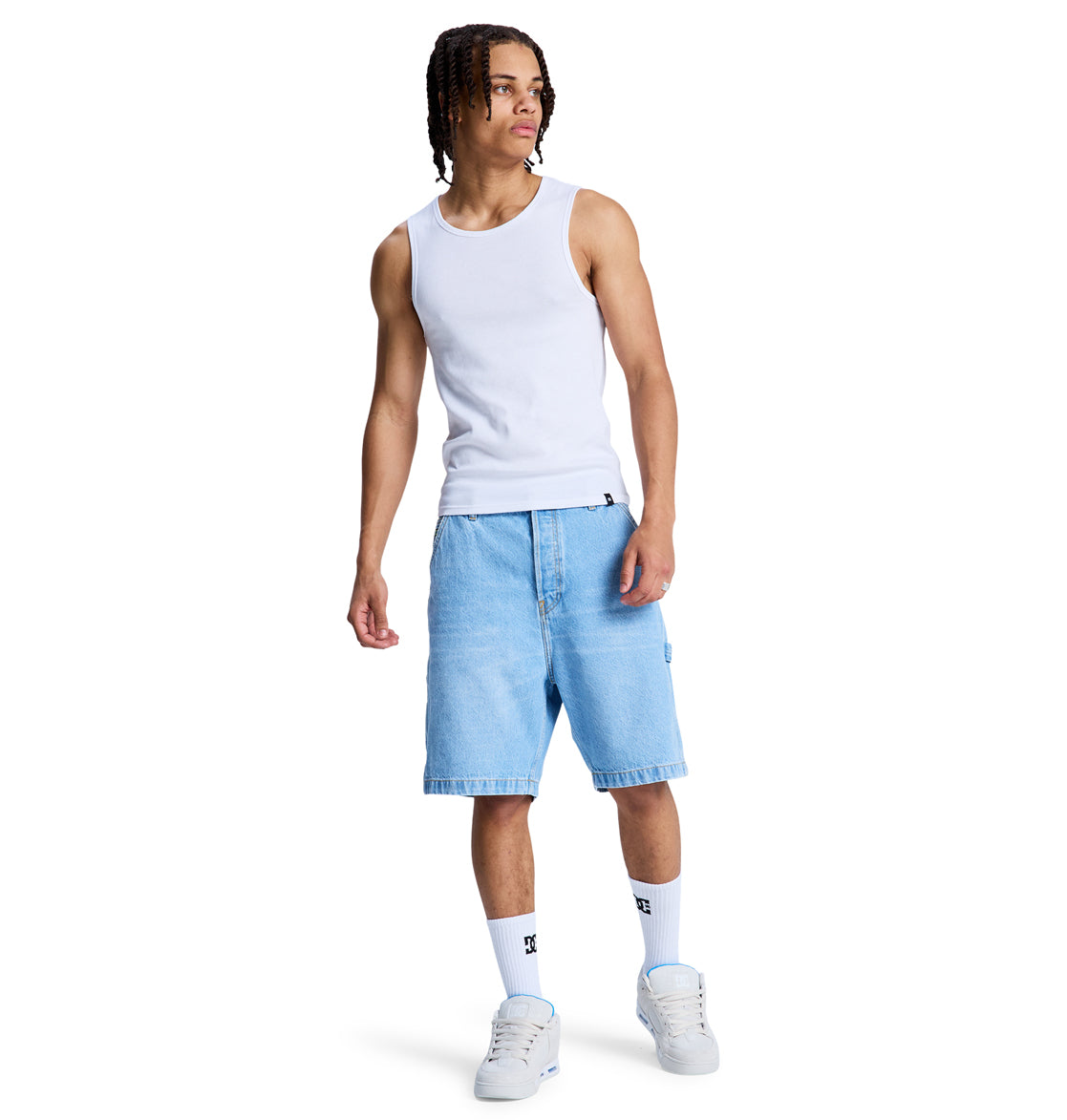 A person wearing a white tank top, Reebok Baggy Denim Carpenter Short in light blue, white socks, and white sneakers stands against a plain white background.