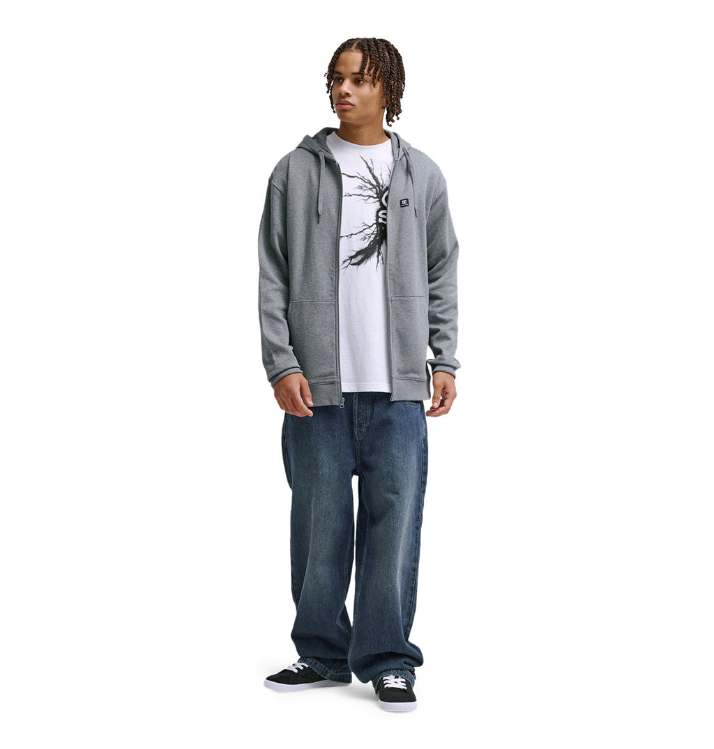 A person stands facing forward, wearing the Reebok Lorion Zh gray zip-up hoodie over a white graphic t-shirt, loose dark blue jeans, and black sneakers against a plain white background.