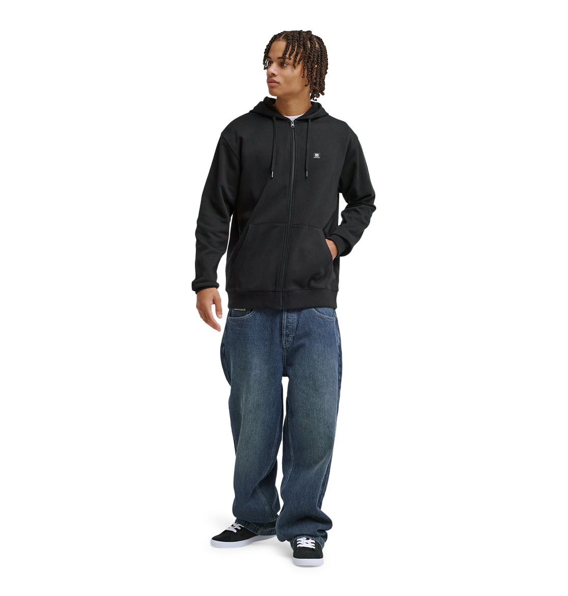 A person with short, curly hair wears a black Reebok Lorion Zh zip-up hoodie, loose blue jeans, and black sneakers while standing against a plain white background.