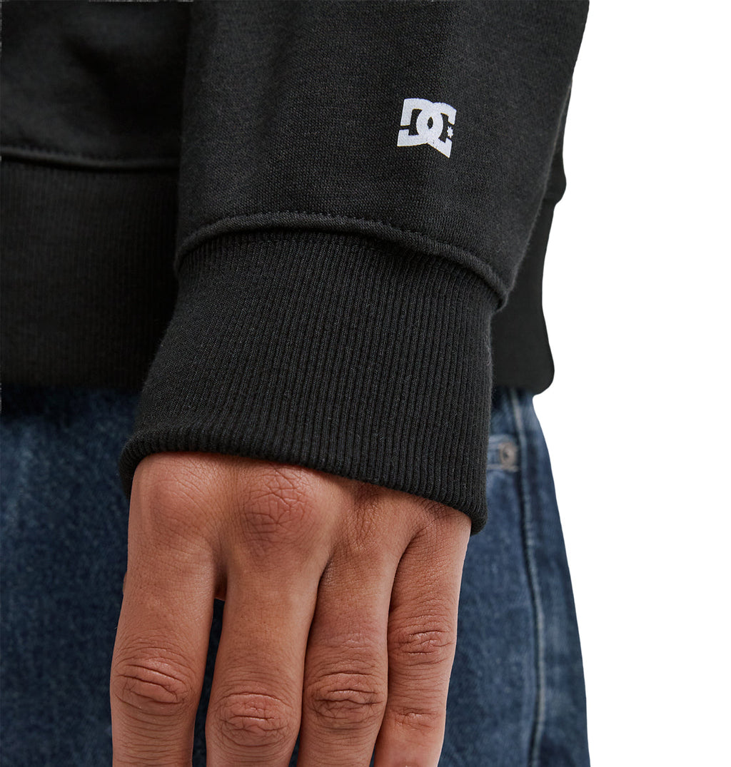 A close-up of a person's hand resting on blue jeans, wearing a black Reebok Lorion Zh sweatshirt with a small white logo on the sleeve cuff.