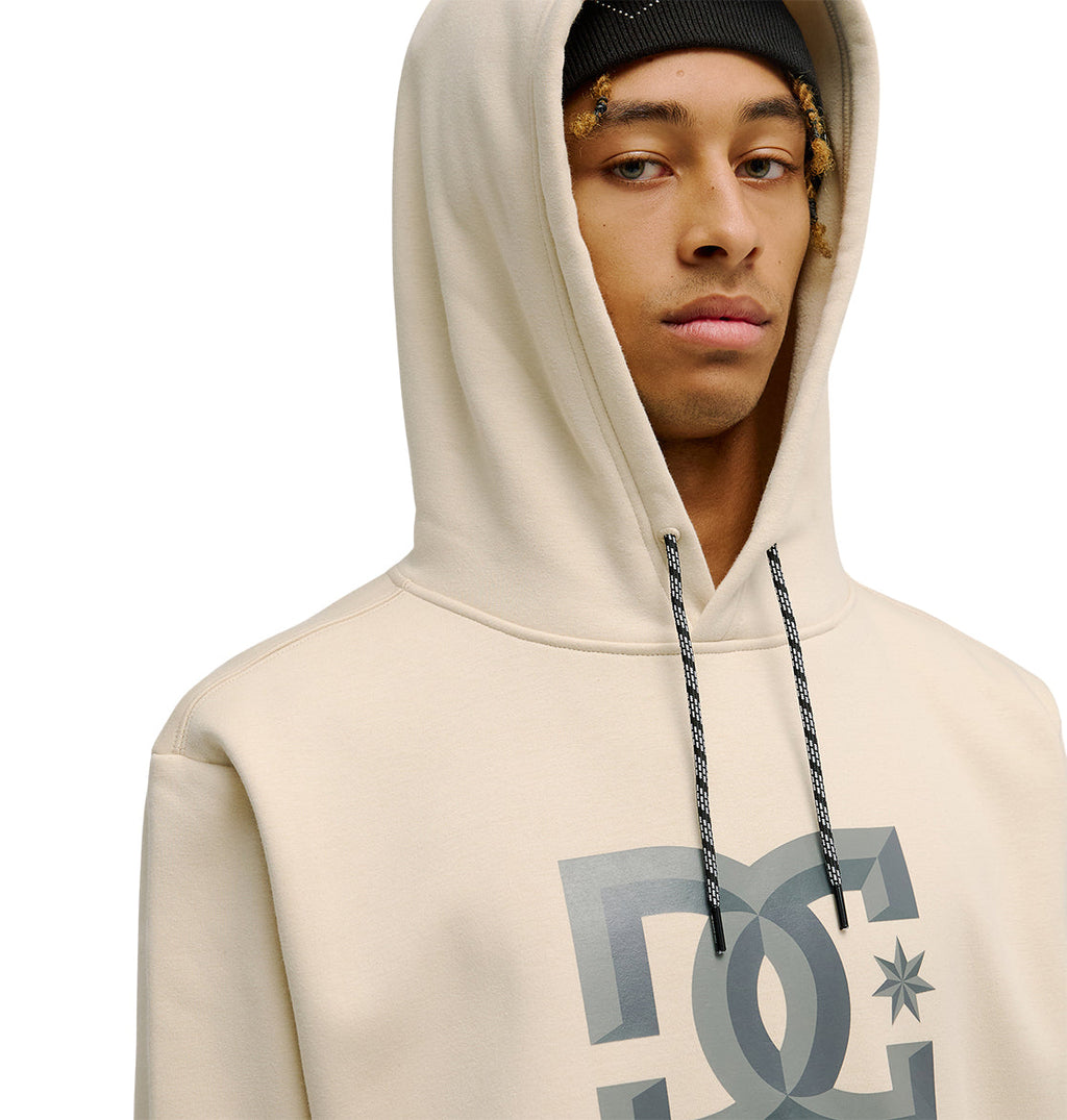 A person wearing the DC Shoes Snowstar beige hoodie with a large DC logo and a black beanie looks at the camera against a plain white background, highlighting both weather protection and a comfortable fit.