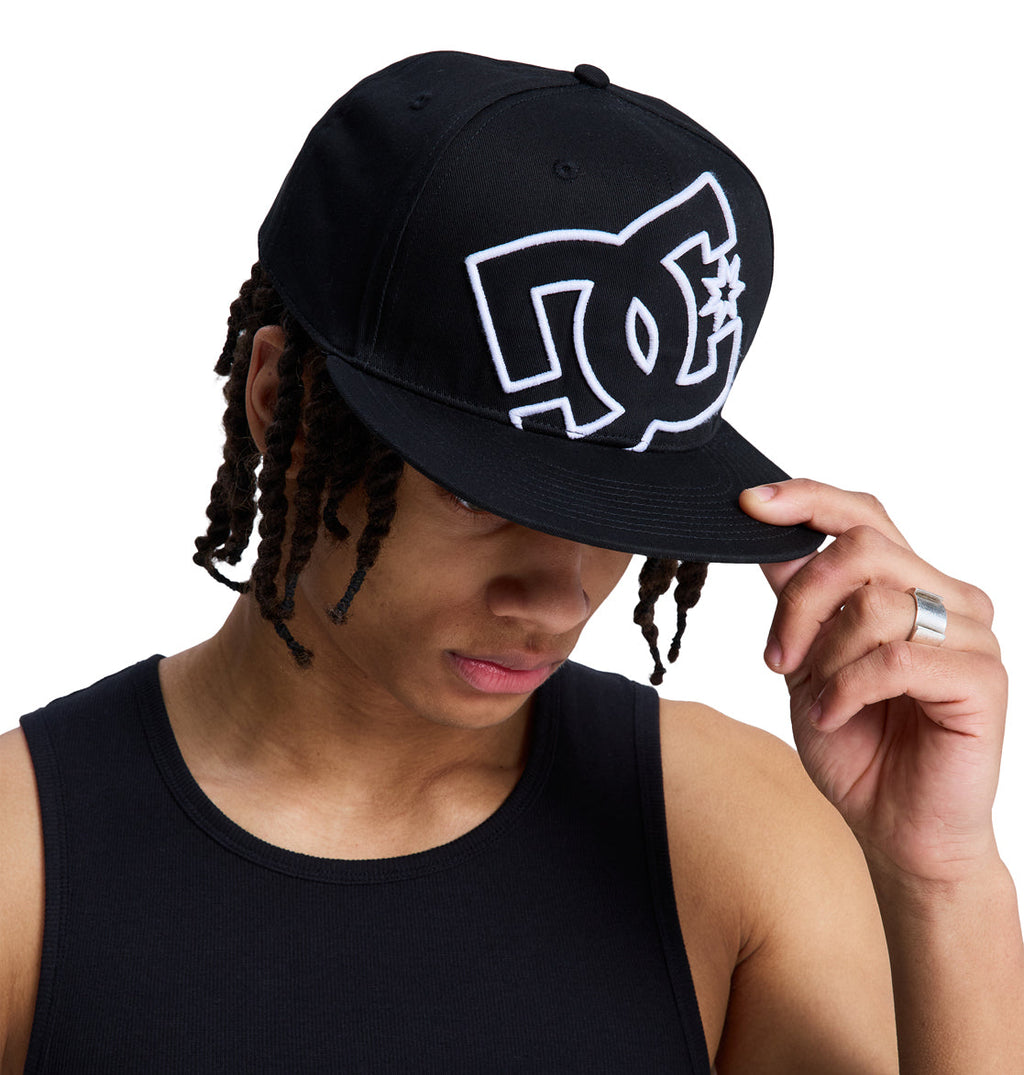 A person in a black tank top and a black Reebok DC Double That Cap tilts the brim downward, partially covering their face. The background is white.