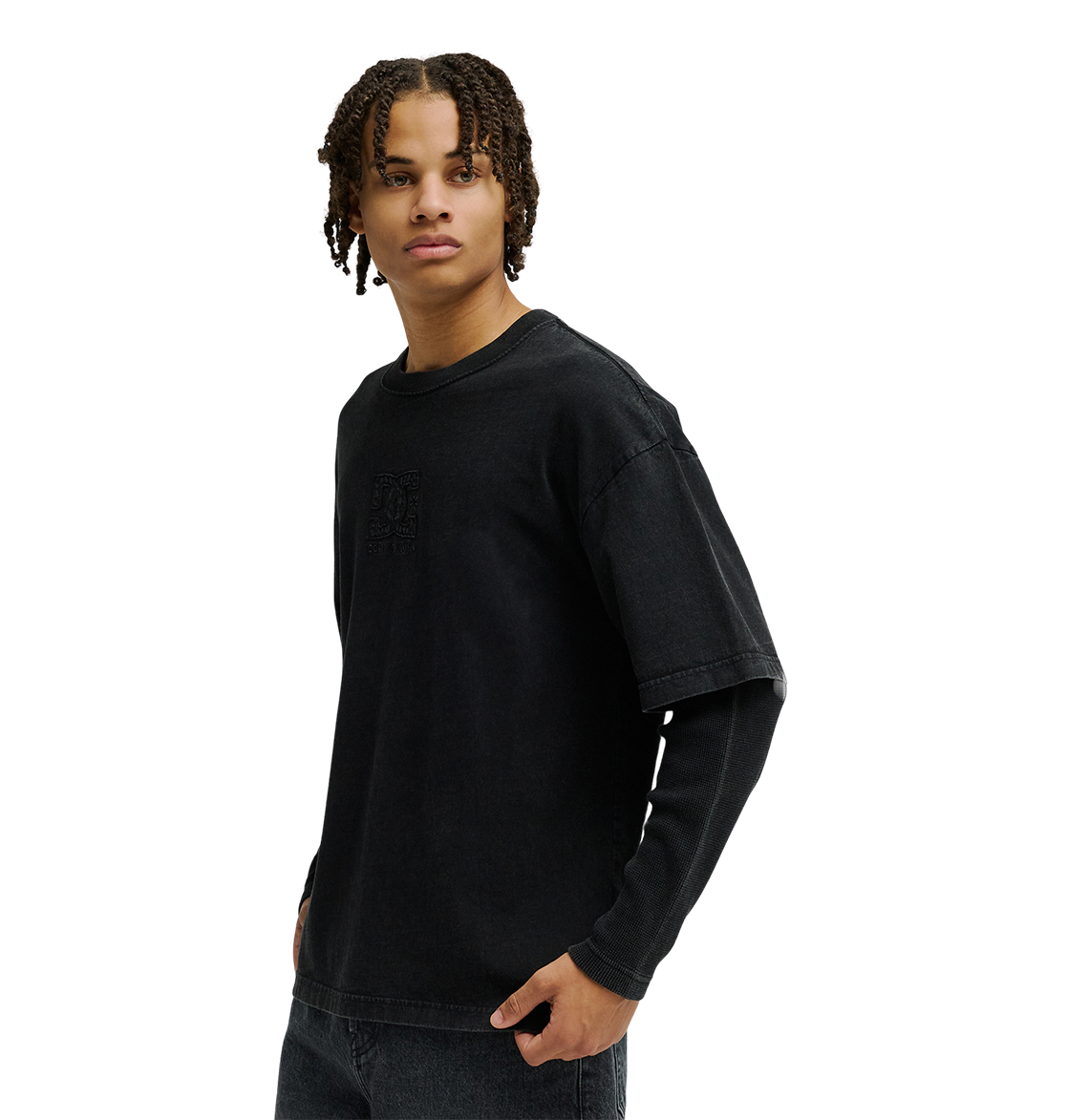 A person with short, curly hair stands against a plain white background, looking to the side, wearing a loose black T-shirt layered over the Men’s DC Twofer Long Sleeve by DC Shoes and paired with black jeans.