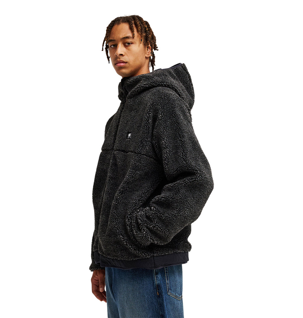 A person wearing the DC Shoes Coldstag Sherpa Reversible jacket—a black, fuzzy fleece design with a hood—paired with blue jeans stands slightly turned against a white background, highlighting its cozy cold-weather comfort.
