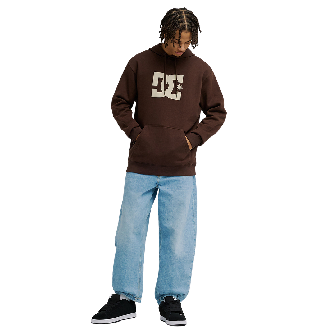 A man wears a brown Men's DC Star Hoodie by DC Shoes, paired with light blue baggy jeans and black sneakers, standing against a plain white background with hands in the hoodie pocket and looking down.