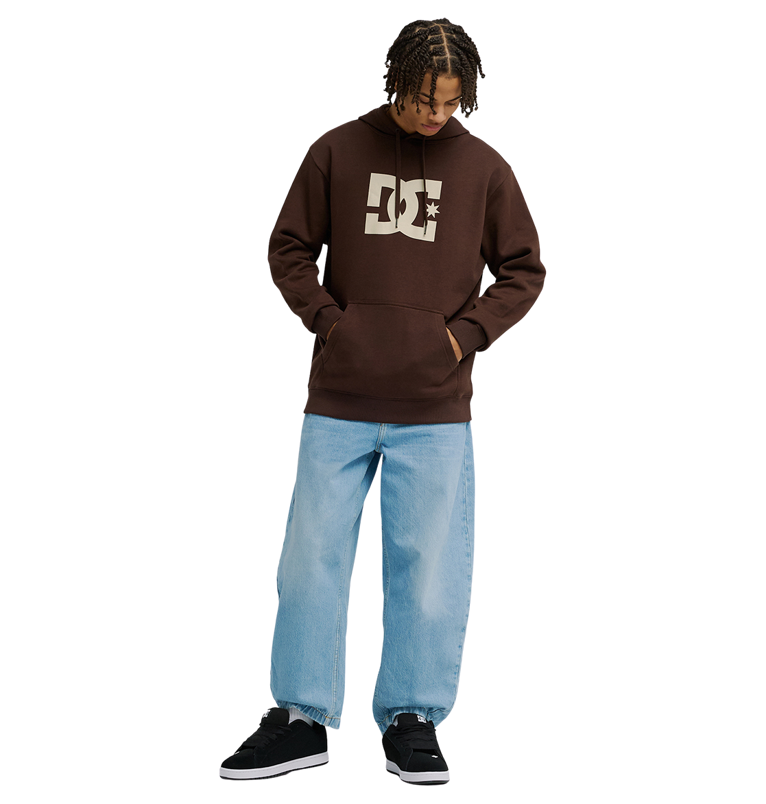 A man wears a brown Men's DC Star Hoodie by DC Shoes, paired with light blue baggy jeans and black sneakers, standing against a plain white background with hands in the hoodie pocket and looking down.