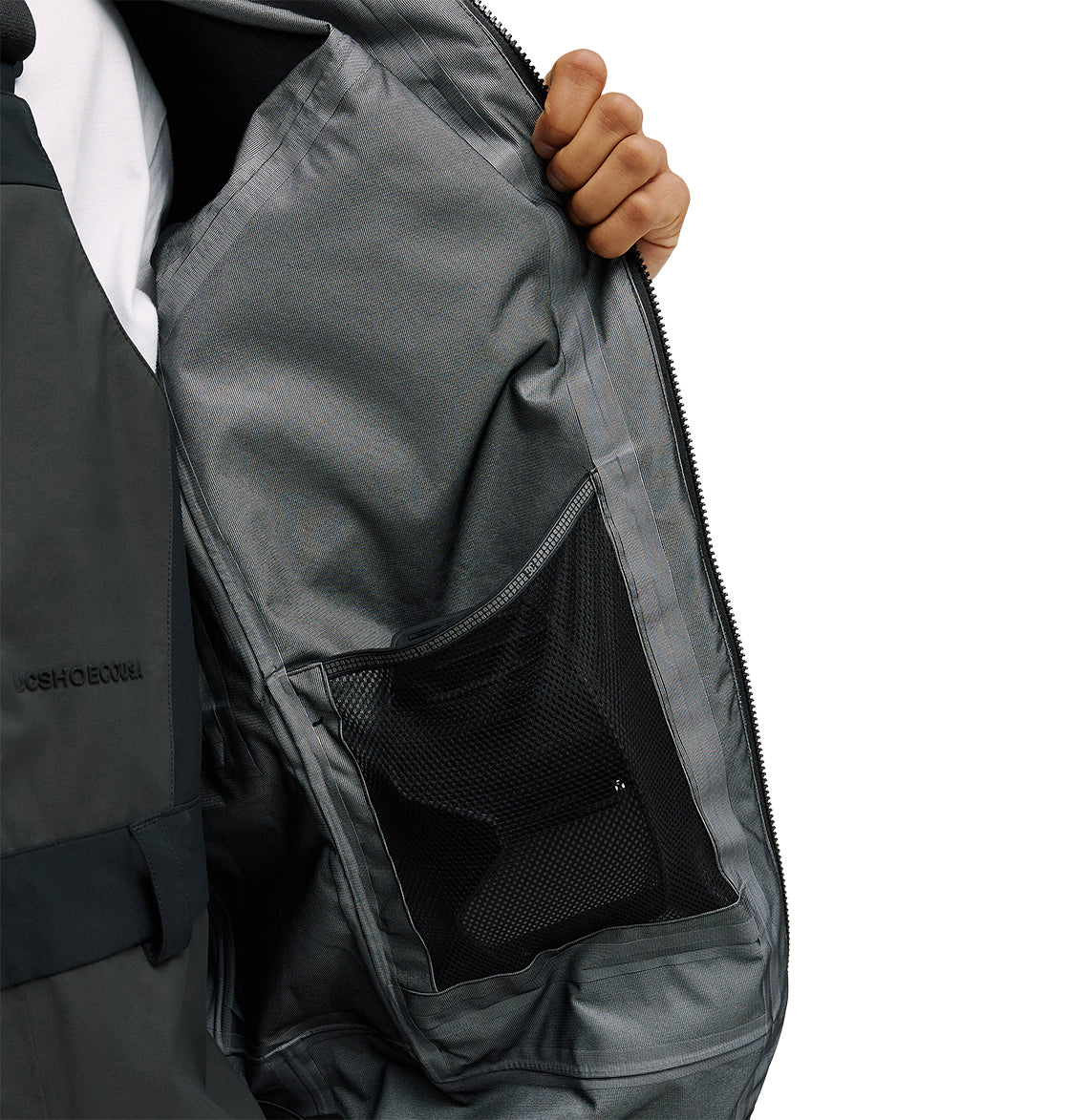A person holds open the DC Shoes Storm Divison 3L Jacket 30K in gray, showcasing its inner lining and spacious black mesh pocket for breathable protection. Only part of the person is visible against a plain white background.