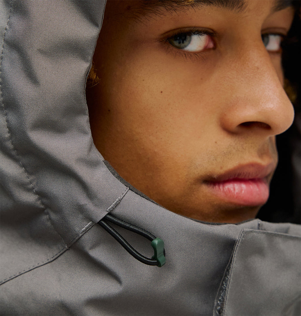 Close-up of a person’s face wearing the Coldsmoke Jacket 10K by DC Shoes—a gray snow jacket featuring a hood with a black drawstring and green toggle for breathable protection, partially revealing their eye and lips.