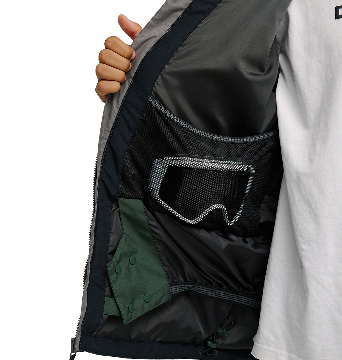 A person opens the DC Shoes Coldsmoke Jacket 10K to reveal a black mesh inner pocket holding ski goggles and a green item, highlighting the jacket’s breathable protection tailored for peak snow performance.