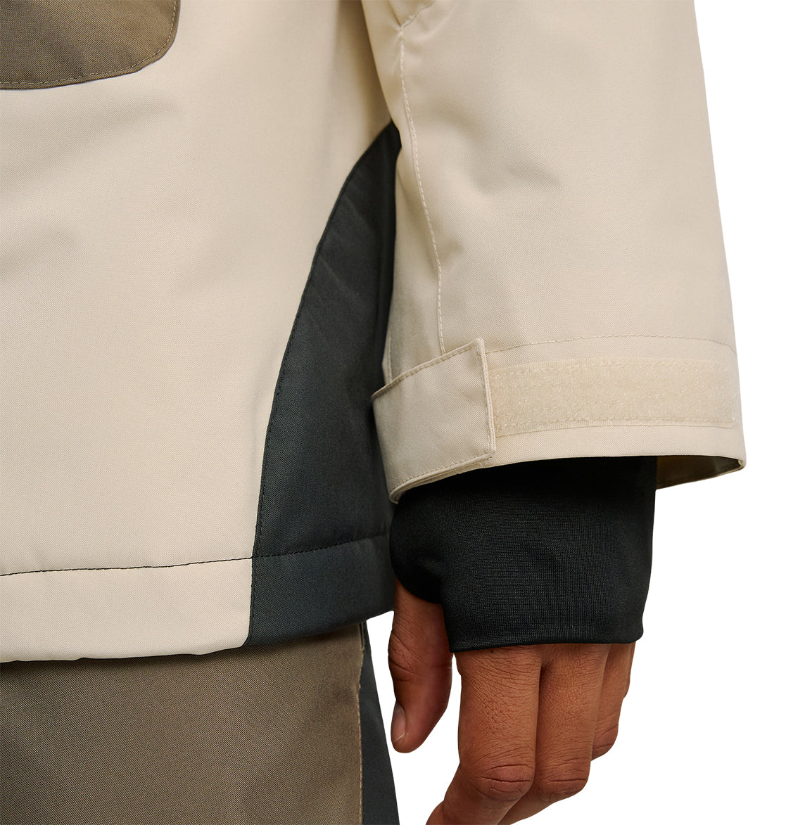 A close-up of a person wearing the DC Shoes Coldsmoke Jacket 10K, featuring color-blocked beige, black, and gray panels, highlights snow performance details like a velcro cuff and a black inner sleeve partially covering the hand.