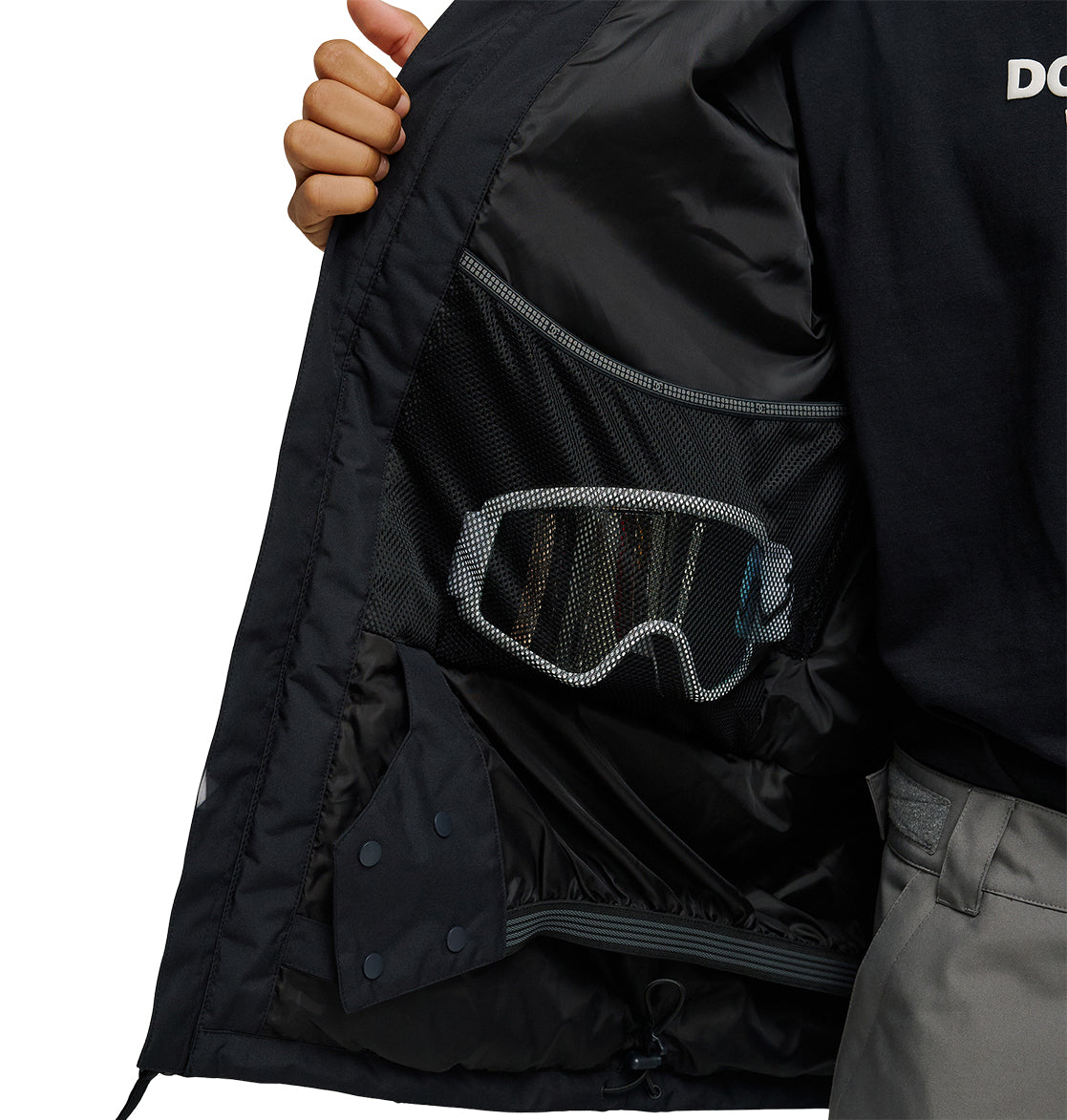A person opens the black DC Shoes Basis Jacket 10K to reveal a mesh inner pocket holding a pair of ski goggles.