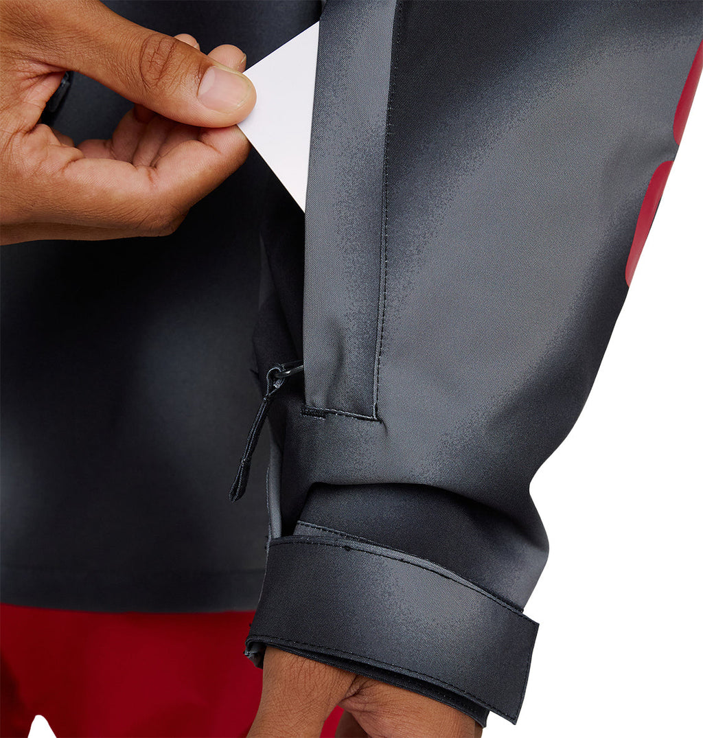 A hand inserts a white card into a small zippered pocket on the sleeve of the DC Shoes Basis Jacket 10K, with red pants visible below.