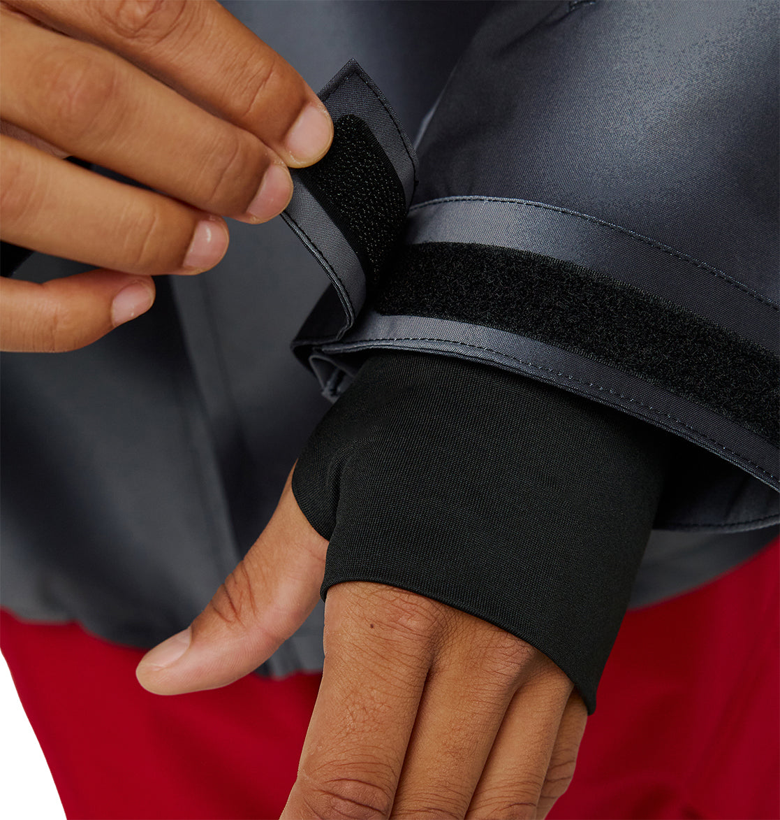 A person adjusts the Velcro cuff of a dark gray DC Shoes Basis Jacket 10K insulated sleeve, revealing a black thumbhole sleeve underneath. Red pants are partially visible.