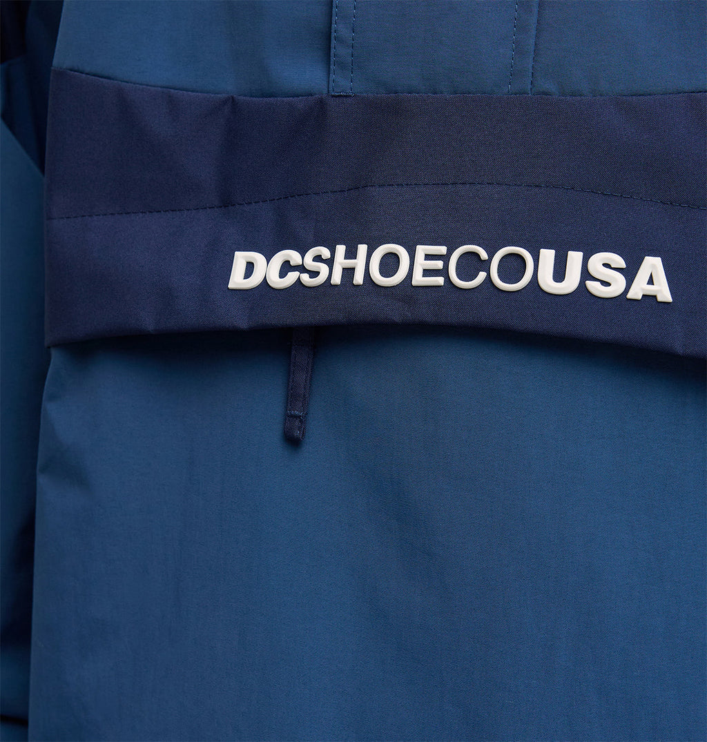Close-up of the DC Shoes Mountain Lab Anorak 10K, a blue fabric jacket with a dark blue stripe and white raised DCSHOECOUSA text, designed for breathable protection and enhanced snow performance.