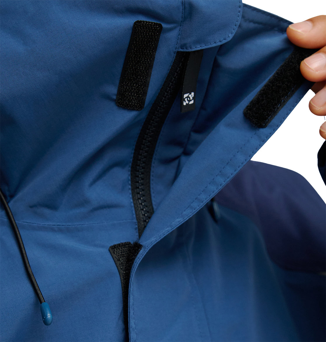 A close-up of a person fastening the hood of the DC Shoes Mountain Lab Anorak 10K using a Velcro strip, with the black zipper and drawstring also visible, highlights its breathable protection for enhanced snow performance.