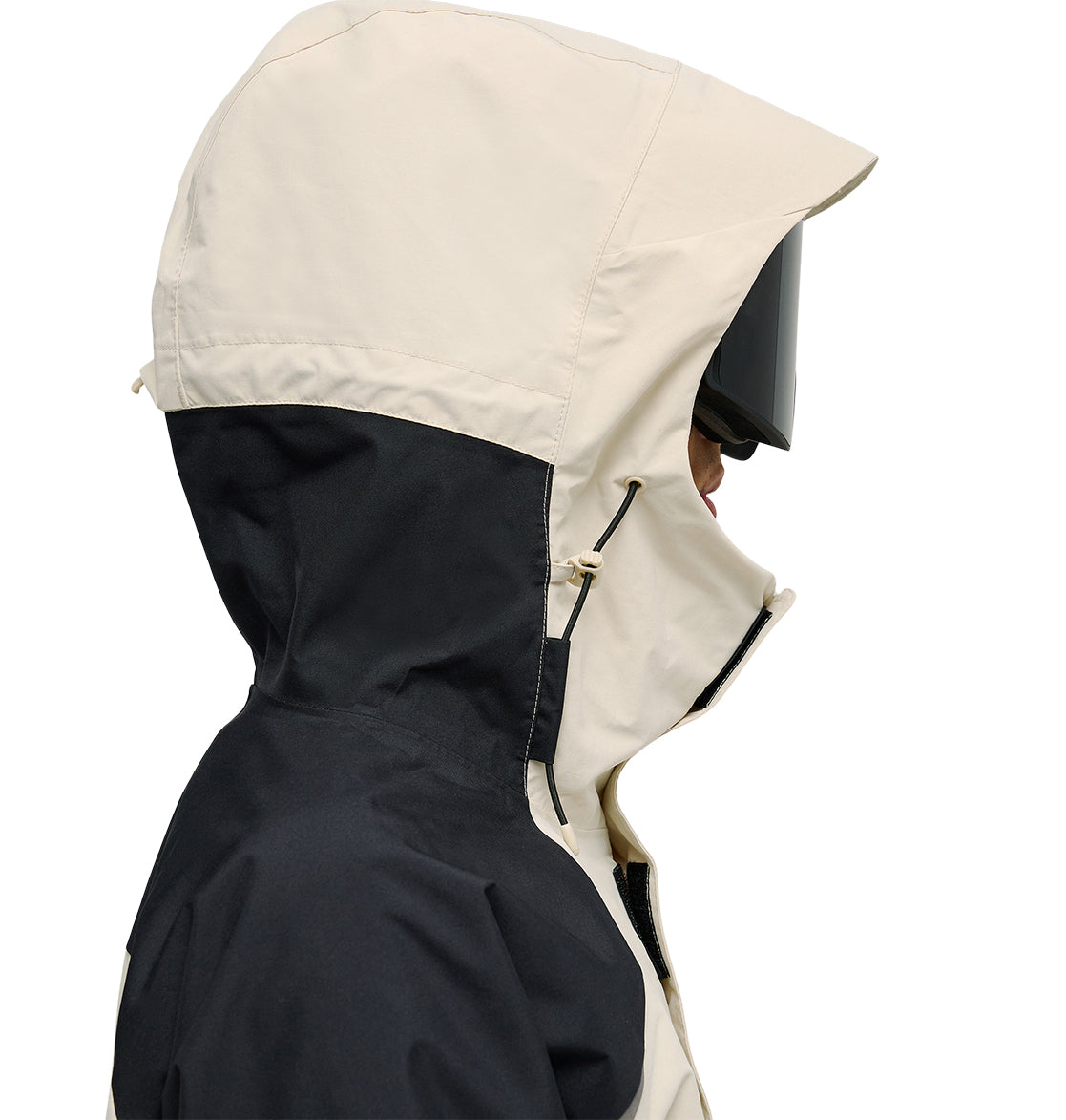 A person in the DC Shoes Mountain Lab Anorak 10K—a beige and black hooded snow jacket—wears dark ski goggles, photographed from the side against a white background, showcasing its breathable protection for winter adventures.
