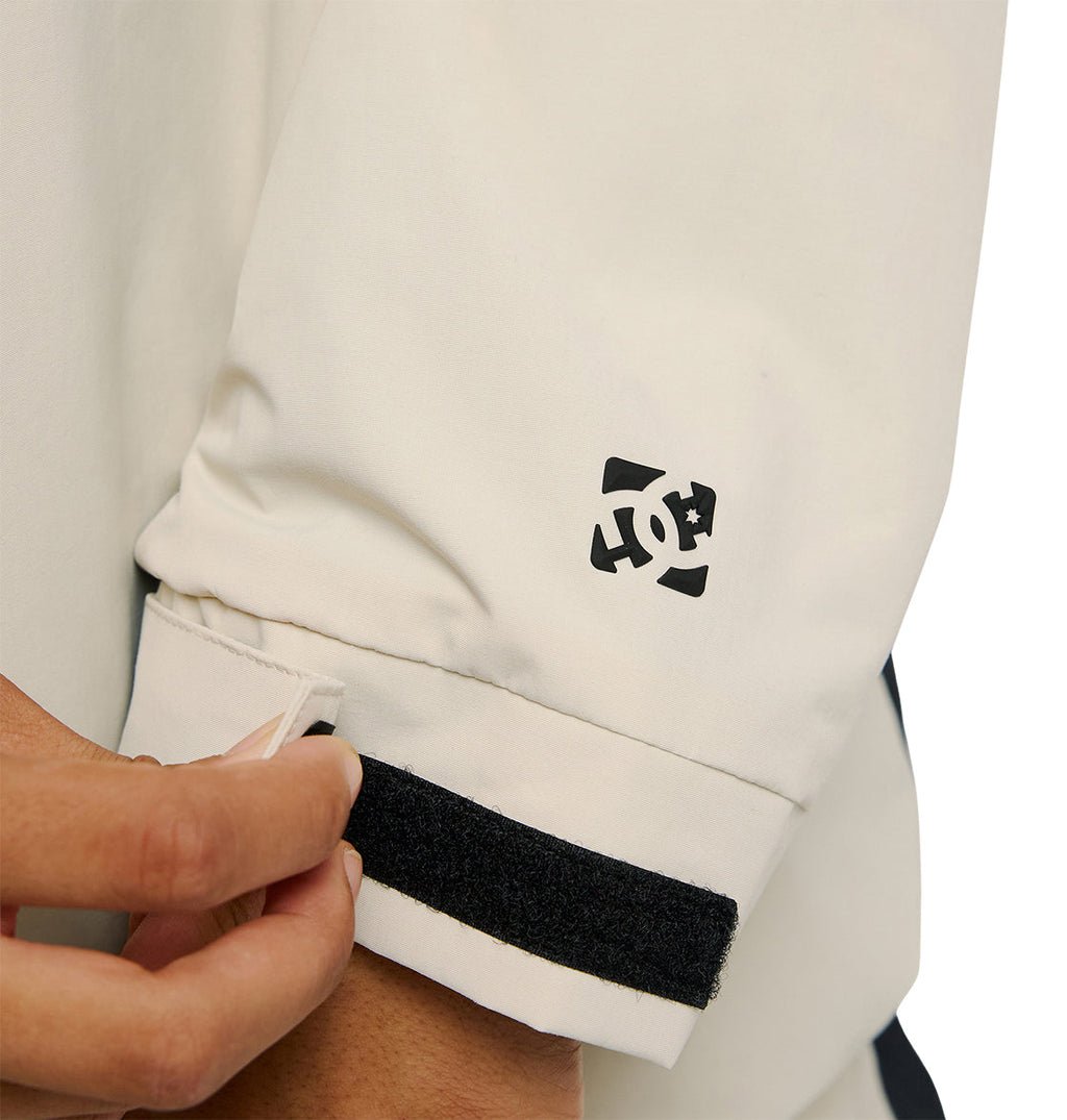 A person adjusts the black Velcro strap on the cuff of a white Mountain Lab Anorak 10K jacket by DC Shoes. Designed for snow performance, this jacket features a small black DC Shoes logo printed near the wrist.
