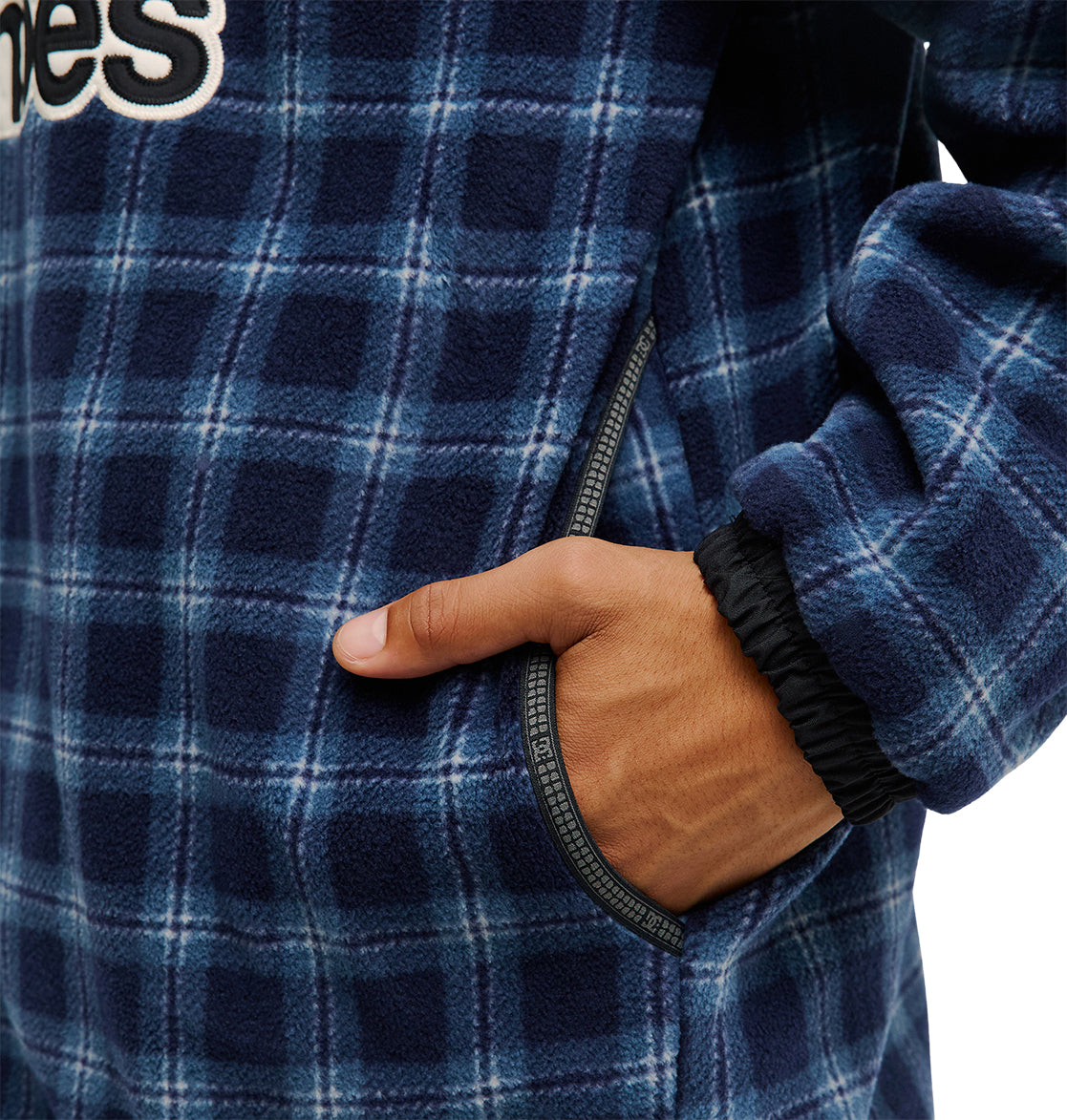 A person wearing the DC Shoes Park Reversible Anorak in blue plaid fleece, featuring black elastic cuffs, has their right hand in a zippered side pocket. This stylish jacket delivers excellent snow performance and reliable waterproofing. Only the torso and arm are visible.