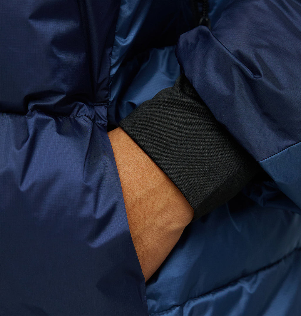 A close-up of a person’s hand in the pocket of the DC Shoes Powder Puffer 10K, a shiny, dark blue jacket designed for superior insulation, with the sleeve of a black shirt visible underneath.