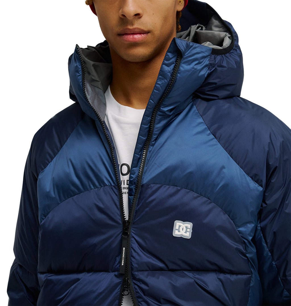 A person wearing the DC Shoes Powder Puffer 10K jacket in blue, partially unzipped to reveal a white T-shirt underneath. Featuring the DC logo on the chest, this hooded puffer’s advanced insulation ensures warmth in chilly conditions. Only the lower half of their face is visible.
