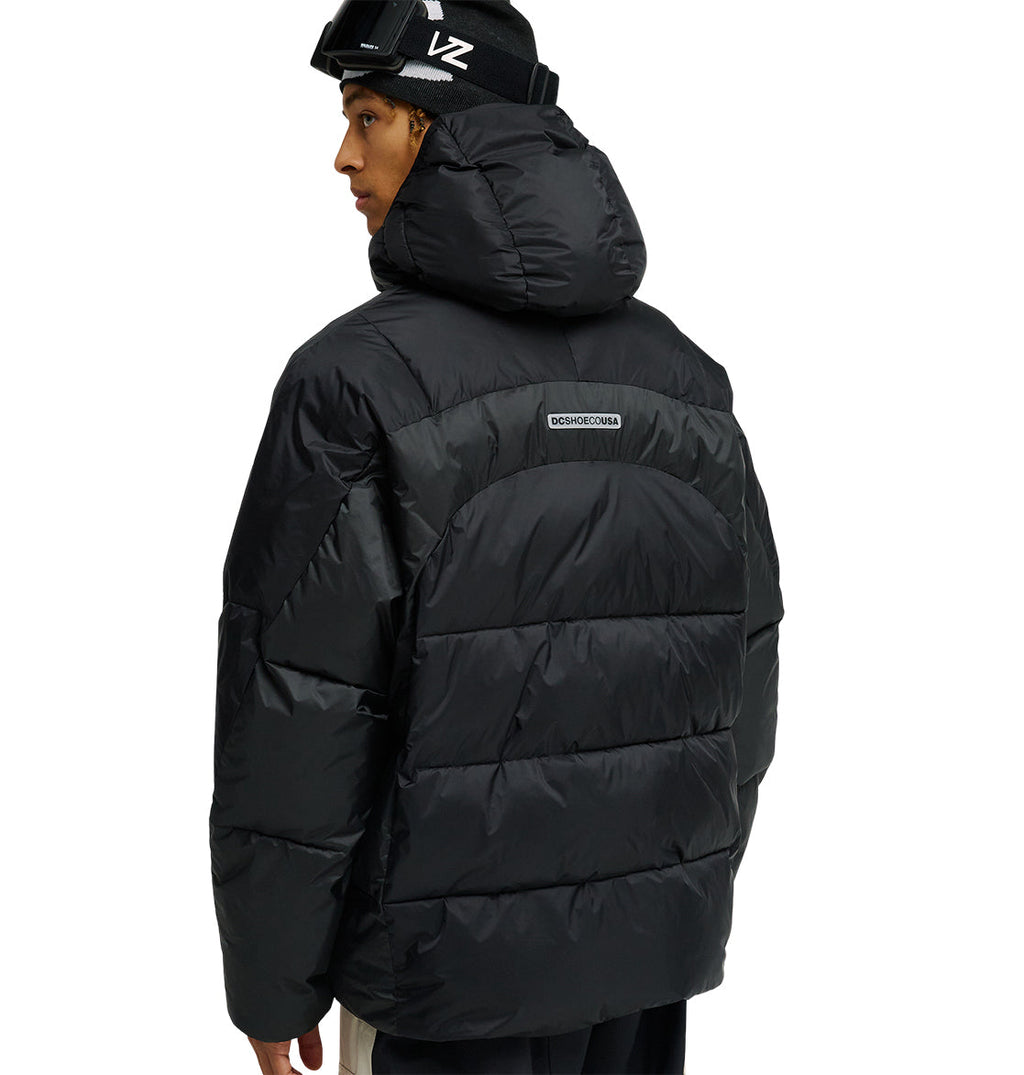 A person wearing the DC Shoes Powder Puffer 10K—a black hooded jacket designed for top snow performance—stands facing away, glancing over their shoulder. The jacket features a small rectangular logo on the back and delivers breathable, element-resistant protection.