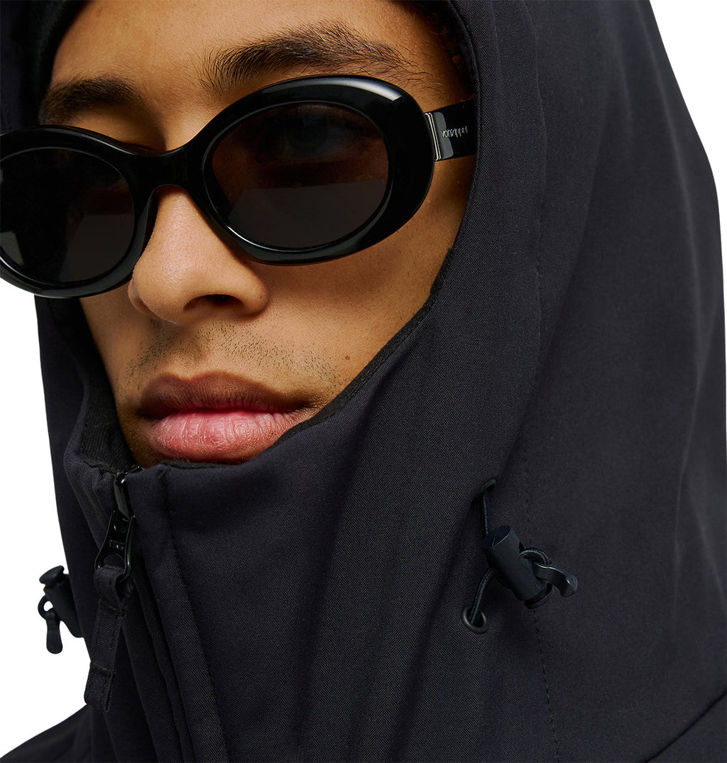 A person wearing the DC Shoes Spectrum Softshell Jacket—a dark, hooded jacket offering waterproof and breathable protection—zipped up to the chin. The look is finished with black oval sunglasses against a white background.