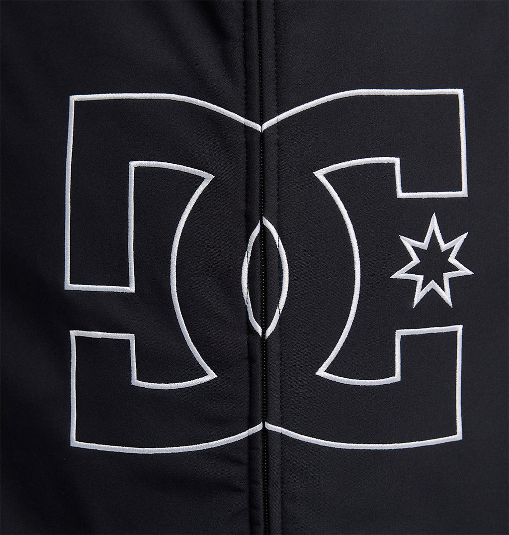 A prominent white DC Shoes logo, outlined in white, is centered on a black fabric background featuring visible vertical stitching—a design element characteristic of the Spectrum Softshell Jacket by DC Shoes, renowned for its waterproof and breathable protection.