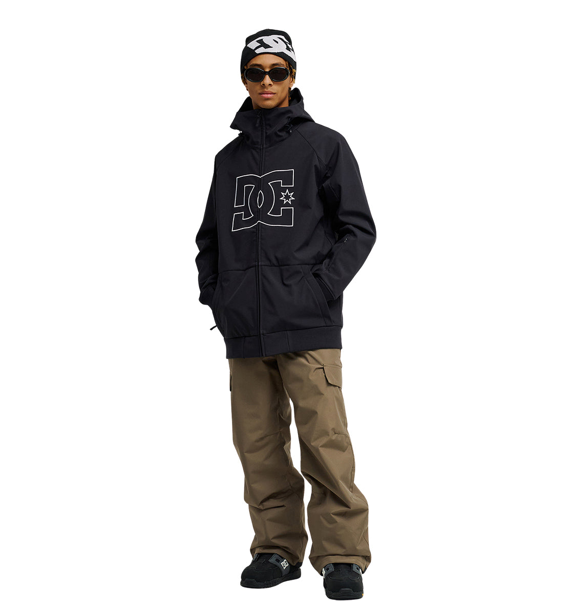 A person stands with hands in pockets against a white background, wearing the DC Shoes Spectrum Softshell Jacket—a black style featuring a large DC logo—paired with khaki cargo pants, black shoes, dark sunglasses, and a black-and-white beanie. The Spectrum Softshell Jacket offers breathable protection for comfort and style in any setting.