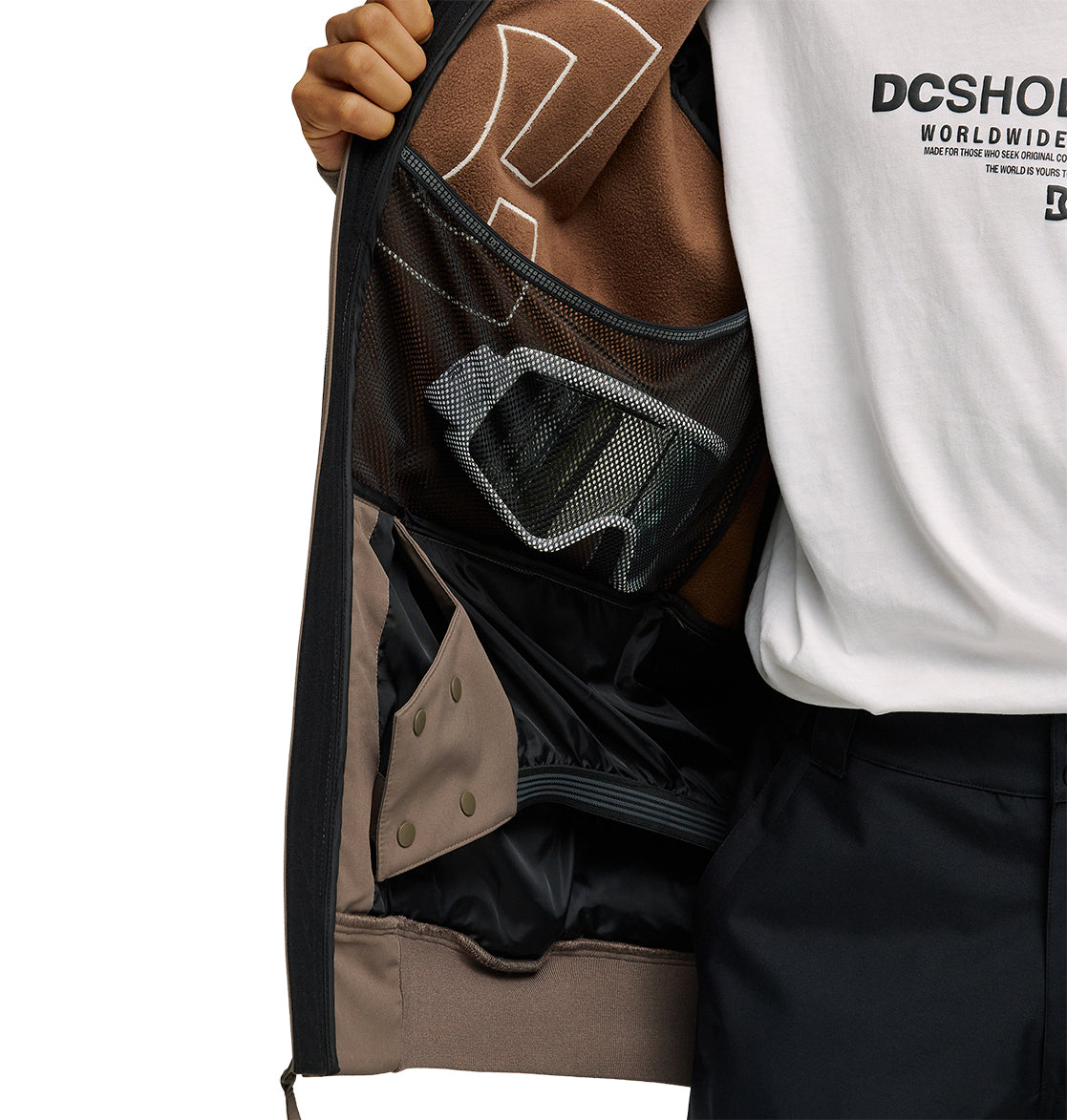 A person opens their DC Shoes Spectrum Softshell Jacket to reveal the inner pocket and mesh compartments, with ski goggles stored inside. The person is wearing a white DC Shoes Worldwide t-shirt and black pants.