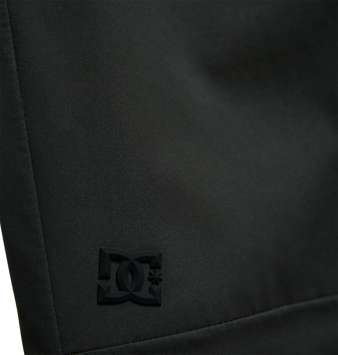 Close-up of the Storm Division 3L Bib 30K by DC Shoes, featuring black fabric with a subtle, embossed DC Shoes logo in the lower left corner. The smooth texture and visible stitching along the left edge showcase its breathable protection—perfect for all-weather comfort and performance.
