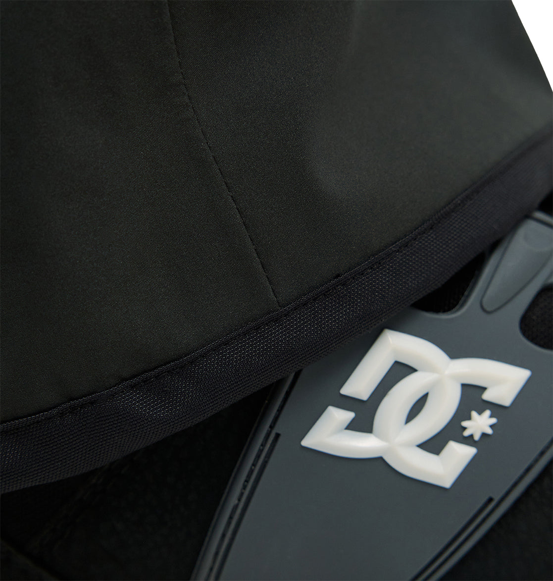 Close-up of the black fabric and seam of the Storm Division 3L Bib 30K by DC Shoes, partially covering a gray section with a raised white DC Shoes logo—highlighting materials engineered for snow performance and breathable protection.