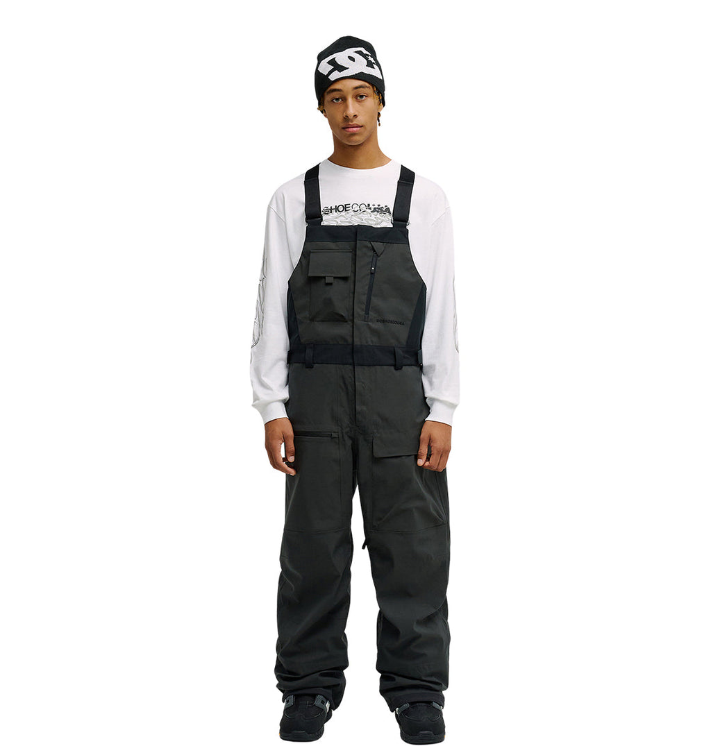 A person stands facing forward with hands at their sides against a plain white background, wearing the DC Shoes Storm Division 3L Bib 30K over dark attire, a white long-sleeve shirt with text, and a black beanie—showcasing this bib’s breathable protective design.