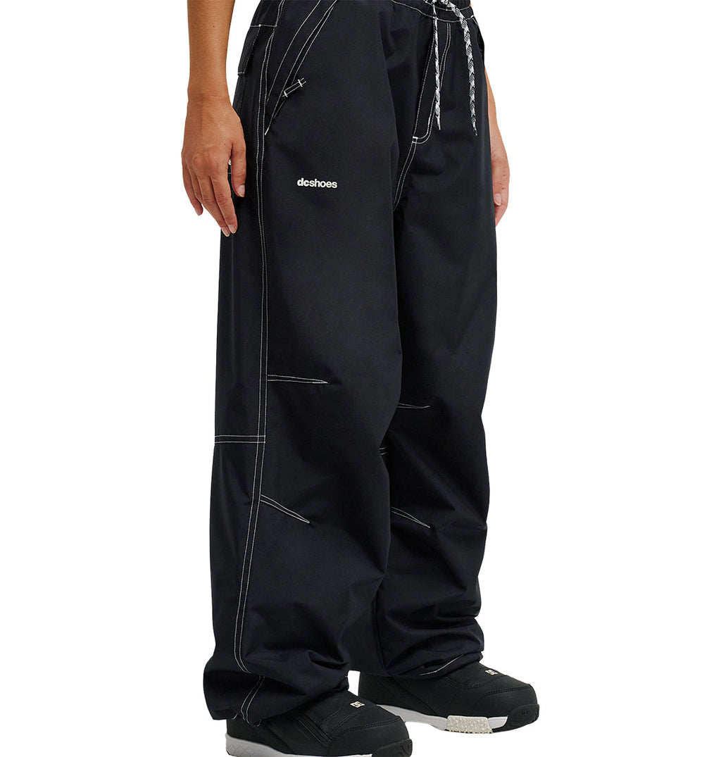A person is seen from the waist down wearing loose-fitting black Trackstar Pants 10K by DC Shoes, featuring white stitching, a black-and-white drawstring, and “DC Shoes” printed on the left thigh. The pants are designed for enhanced snow performance and paired with black shoes.