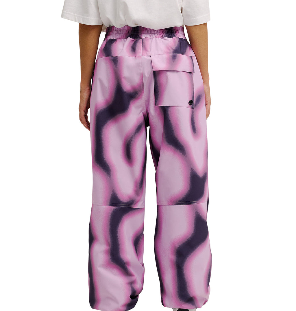 A person is seen from the back wearing DC Shoes Trackstar Pants 10K—loose, high-waisted waterproof pants with a pink and black wavy pattern. The pants provide breathable protection, feature a large right-side pocket, and have elastic at the waist and ankles.